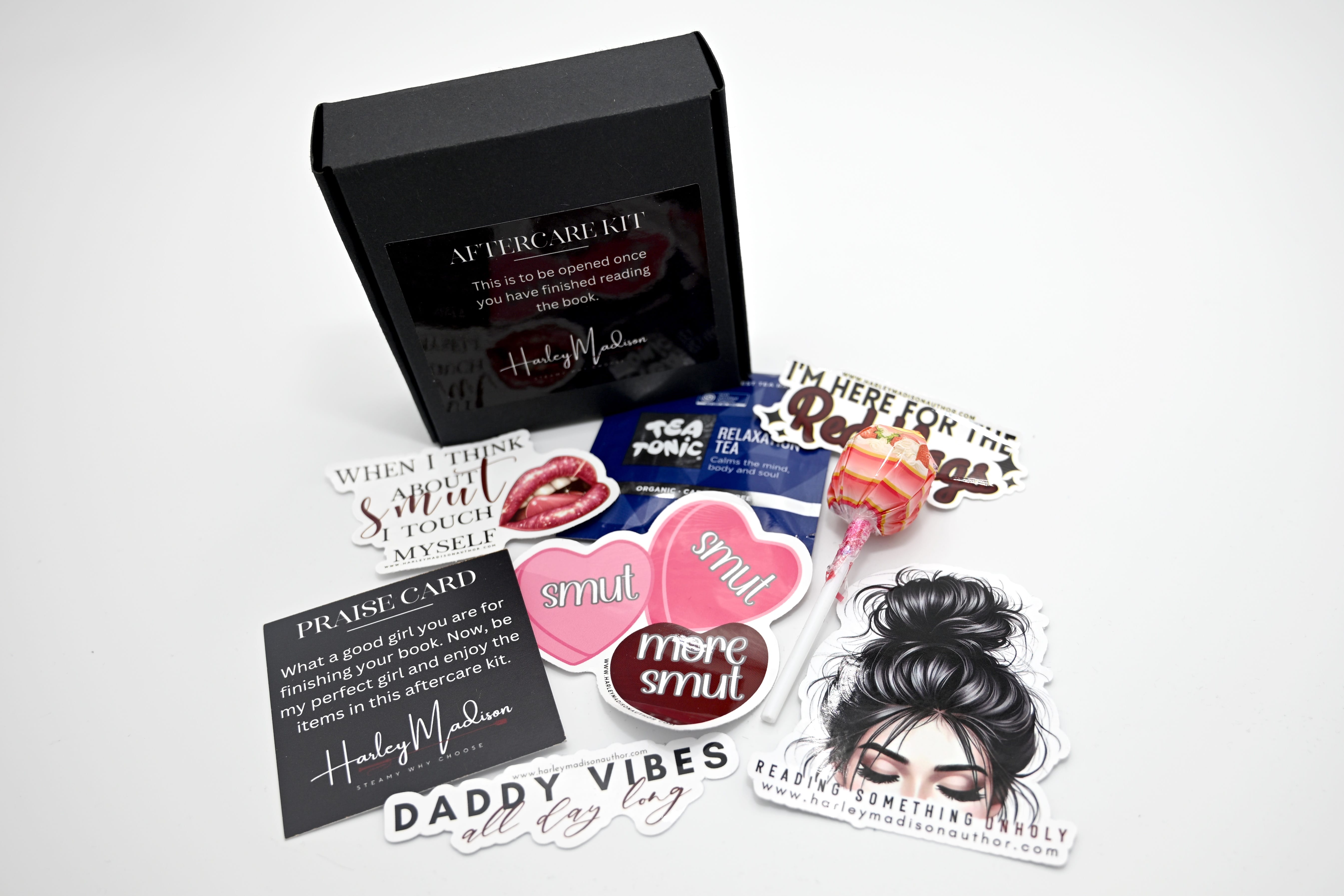 An open "Aftercare Kit" box by author Harley Madison, surrounded by themed items including a "Praise Card," a pink lollipop, a tea packet, and several stickers with romantic or "smut" themed text.